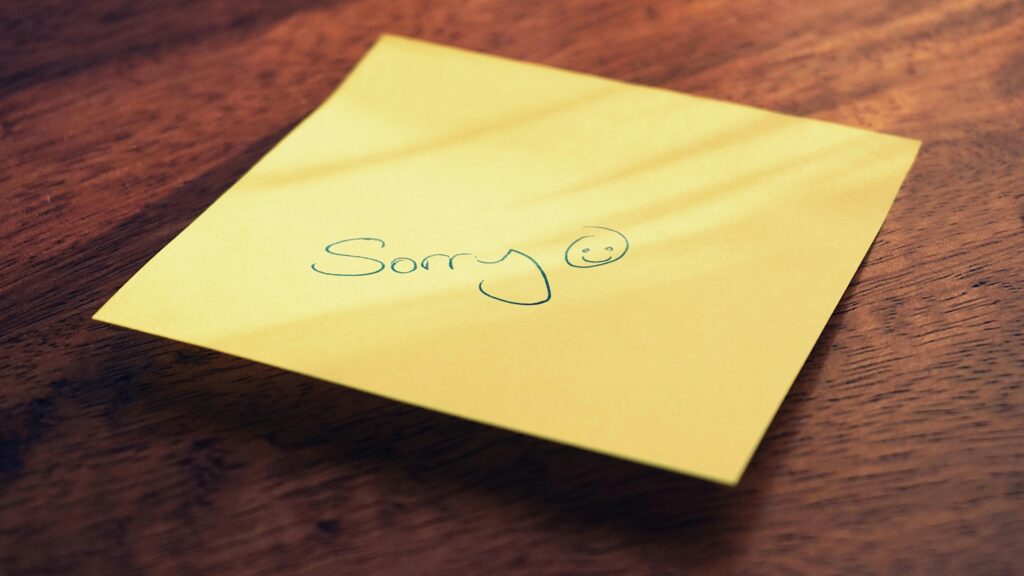 A yellow post it note with the word "Sorry" on a brown wood table.