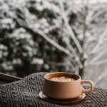 An image of a cup of coffee on a blanket in front of a view of trees with snow.